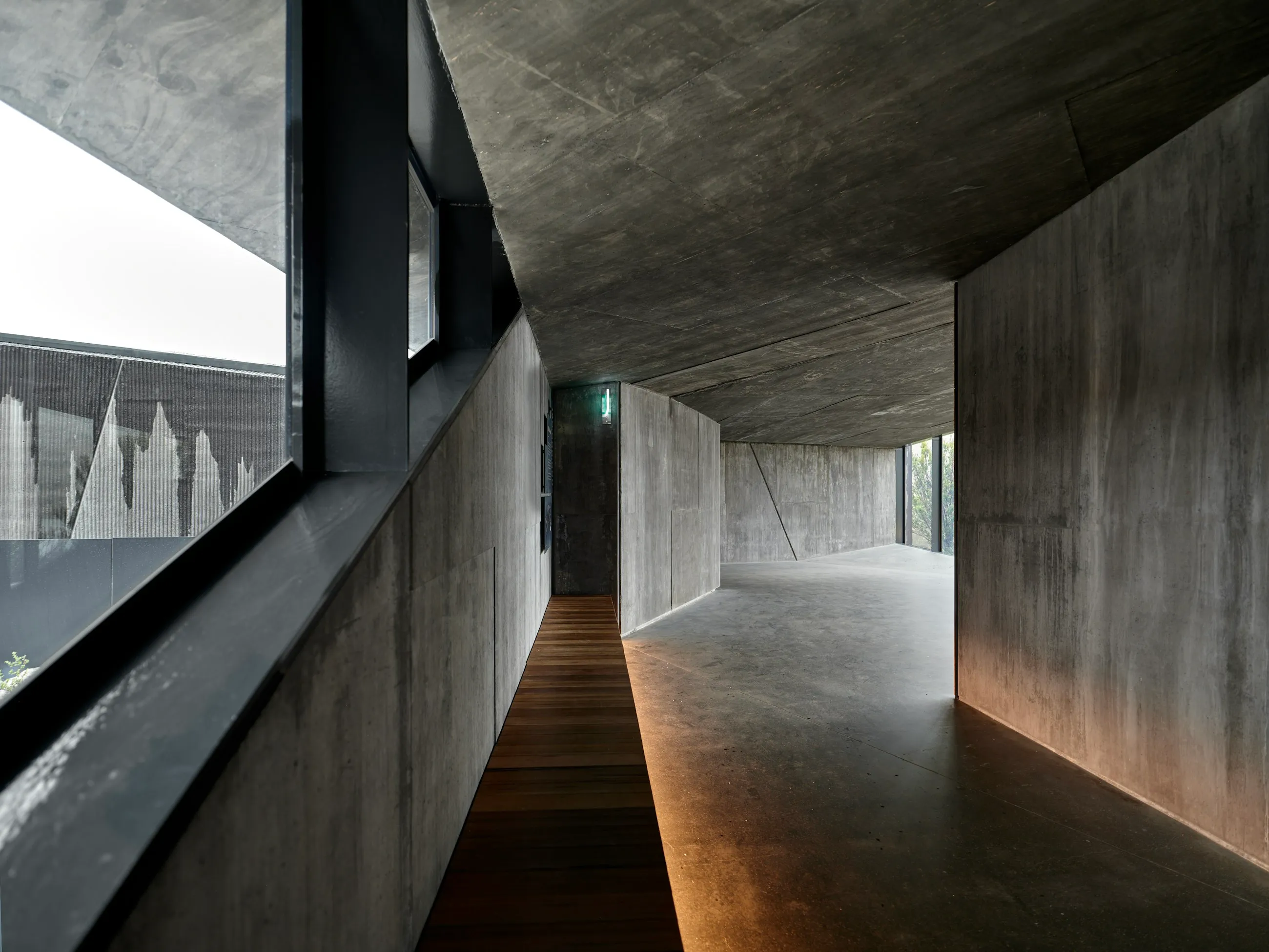 A dark concrete corridor with daylight falling along a timber floor toward an opening.