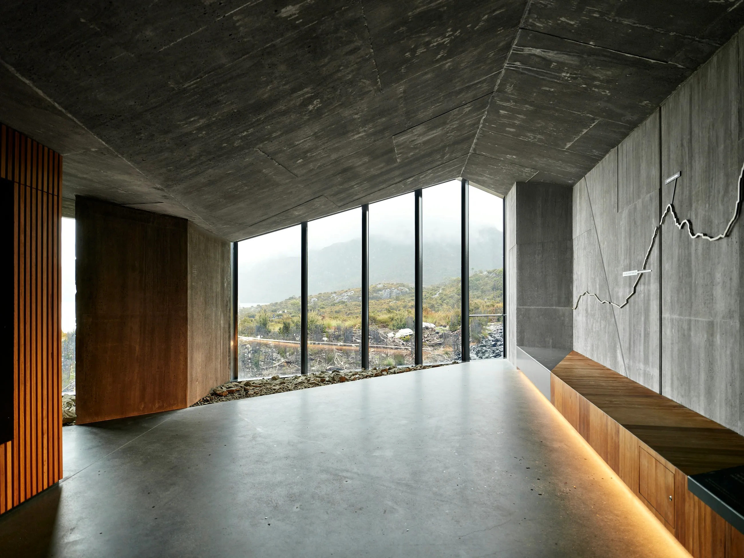 A concrete and timber room opening through full-height glazing onto a misty mountain landscape, raking light along a timber bench.