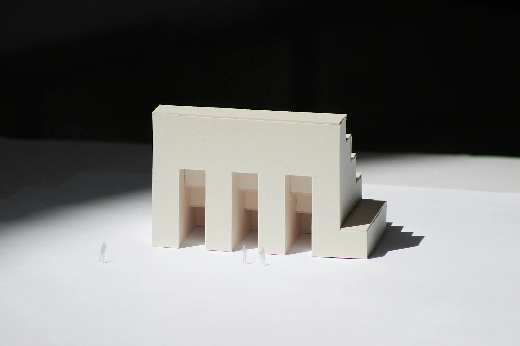 A white card architectural model lit by hard side light, with three tiny scale figures.